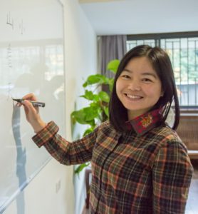 A teacher smiles over her shoulder while writing on a whiteboard by a plant