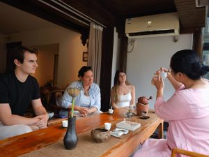 A tea host raises a small cup while three people watch at a long table