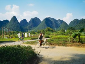 A woman cycles along a farm road below green karst peaks