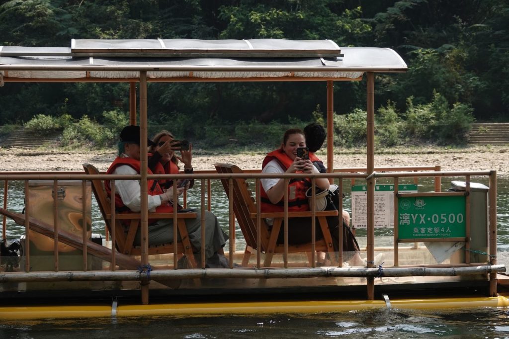 Studenten in orangefarbenen Schwimmwesten fotografieren von einem überdachten Bambusfloß auf dem Fluss aus