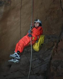 A person in orange coveralls and a helmet rappels down a cave wall