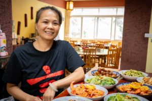 A smiling cook in a CLI T-shirt sits behind bowls of shrimp, pork and greens, with a dining room behind her
