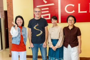 Four people smile in front of a red wall with the 言 CLI logo, beside a column covered in signatures