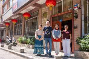 Four people stand outside a brick building's wooden door under red lanterns, house number 31 above
