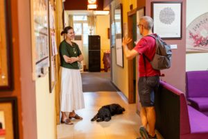 A man with a backpack waves to a smiling woman in a hallway as a black dog lies on the floor