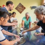 A cook and a group stand around a table folding dumplings from a bowl of green filling