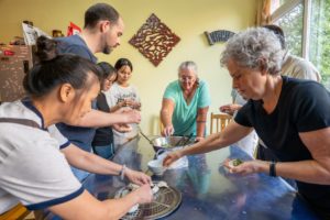 A cook and a group stand around a table folding dumplings from a bowl of green filling