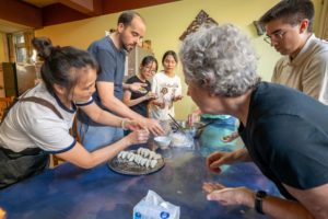 A cook laughs while folding a dumpling as others around the table copy her, a tray of finished dumplings in front