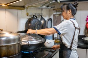A cook in an apron lifts the lid of a wok on a gas stove in a home-style kitchen