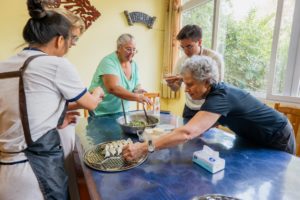 A woman sets a folded dumpling on a steamer tray as others fill wrappers around a blue table