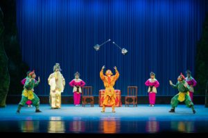 Costumed opera actors on a blue-curtained stage as a performer in gold tosses two maces into the air