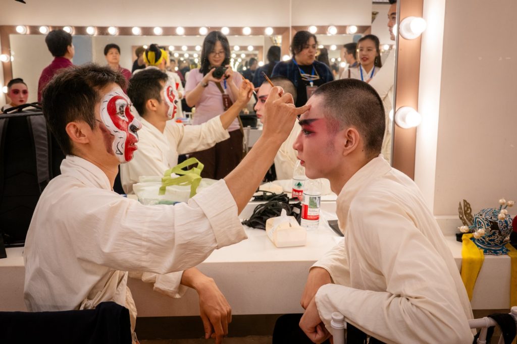 A Guilin Opera performer paints another performer's face at a backstage mirror as onlookers watch