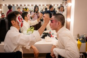 Backstage, a performer in red-and-white face paint draws the eyebrows of another performer at a lit mirror