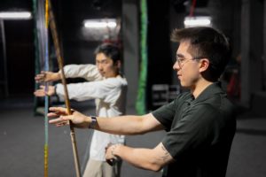 A performer in white shows a man in a dark shirt how to hold a long spear in an opera stance