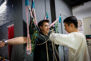A performer ties four embroidered opera back flags onto a smiling man in glasses