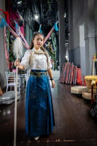A woman in a white blouse and blue skirt wears four embroidered back flags and holds a tasseled spear backstage