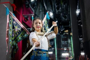 A smiling woman wearing embroidered back flags holds a spear across her body backstage