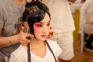 Someone fits a black opera headpiece on a person in pink-and-red stage makeup