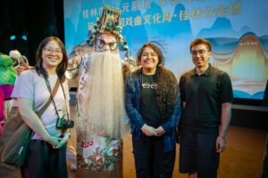 Three smiling people pose on a stage with a performer in an ornate headdress and long white beard