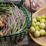 A woven basket of long beans and fresh ginger beside a bamboo tray of round green fruit
