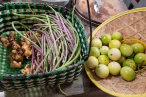 A woven basket of long beans and fresh ginger beside a bamboo tray of round green fruit