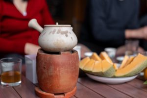 A clay teapot heats on a small terracotta stove beside a plate of sliced melon
