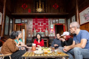Six people sit around a low tea table with melon and fruit in a wooden farmhouse hall hung with red 囍 characters