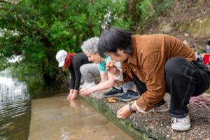 Three women crouch at the water's edge to rinse fresh ginger
