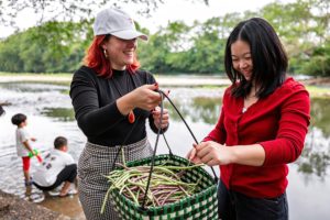 Two women laugh over a basket of long beans by a river where children play