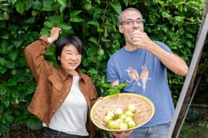 A woman reaches up to a fruit vine as a man bites into a fruit from the basket he holds