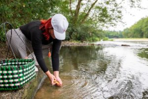 A woman in a white cap leans down to rinse ginger in a clear river beside a woven basket