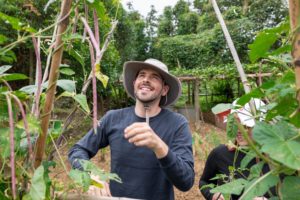 A smiling man in a wide-brimmed hat holds a long bean under a bean trellis