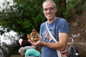 A smiling man in glasses holds a handful of washed ginger by the water as others rinse more behind him