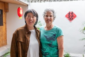 Two smiling women stand together in a white courtyard with a red lantern and a red 囍 sign