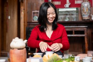 A smiling woman in red arranges small cups at a tea table in a wooden farmhouse hall