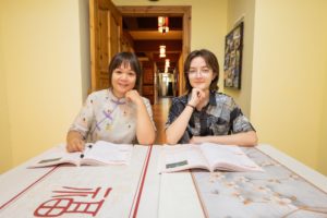 A teacher and a student rest their chins on their hands behind open workbooks, a lantern-lit hallway behind them