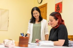 A teacher smiles as her student laughs and writes at a desk under a red 福 poster