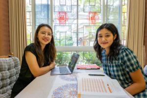 A teacher and a student smile at a table with a laptop and open textbooks by a lattice window with red paper-cuts