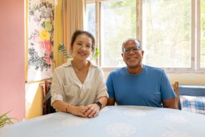 A teacher and a student smile side by side at a table in a bright classroom with a flower painting