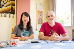 A smiling teacher and student sit side by side behind open textbooks and a mug