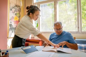 A teacher leans over to point at a textbook page as her student follows, pen in hand, beside a character practice sheet