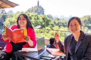 A student holds up a red Chinese textbook beside her laughing teacher at an outdoor café table, a pagoda on a hill behind