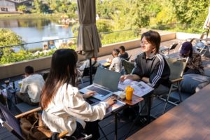 A teacher and a student work at a laptop and printed pages at an outdoor café table above a lake