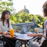 A teacher at a laptop talks with a student across a small outdoor table, a pagoda-topped hill behind them