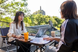 A teacher at a laptop talks with a student across a small outdoor table, a pagoda-topped hill behind them