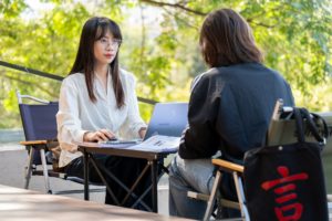 A teacher at a laptop faces a student across a small outdoor table, a black tote with a red 言 on the chair