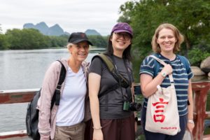 Three smiling women stand at a railing by a river with karst hills behind