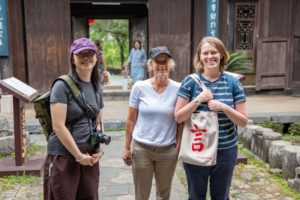 Three smiling women pose on a stone path in front of an old wooden hall