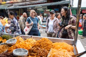 Visitors smile over a market stall piled with trays of fried snacks
