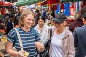 A woman laughs as she holds a half-eaten fruit in a crowded street market, a woman in a black cap smiling beside her
